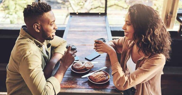 A smiling couple sits at a rustic wooden table in a cafe, holding coffee cups. Pastries on plates are in front of them, specially chosen for their food allergies. A smartphone lies on the table. Sunlight streams through large windows, creating a warm atmosphere perfect for dating with asthma concerns.