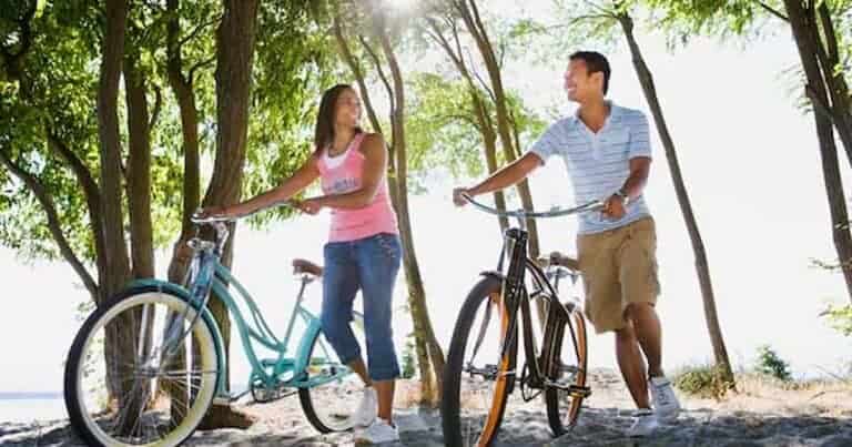 Your Trusted Resource 12 A woman and a man walking with bicycles through a sunlit, wooded area. The woman, in a pink tank top, and the man, in a striped shirt, are smiling at each other. Sunlight filters through the trees, casting dappled shadows on the ground.