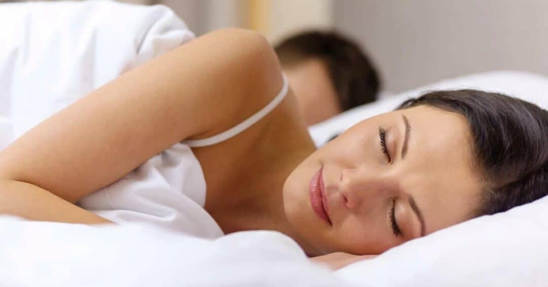 A woman with dark hair is peacefully sleeping on her side in a bed, covered with a white sheet. Another person is partially visible, sleeping in the background. The room has a calm and serene atmosphere.