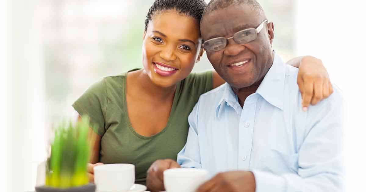 A woman in a green shirt and an older man in a blue shirt and glasses smile while sitting together. They both hold white mugs. There is a small green plant in the foreground.