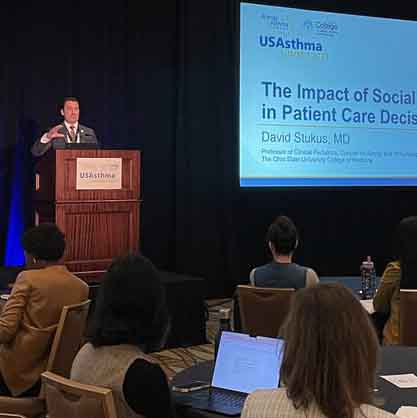 A speaker at a conference stands at a podium delivering a presentation titled "The Impact of Social Media in Patient Care Decisions." The audience is seated, some with laptops open, in a dimly lit room.