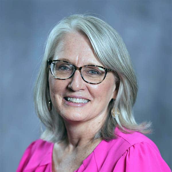 De De Gardner, a woman with glasses and shoulder-length gray hair smiles at the camera. She is wearing a bright pink blouse. The background is a soft, blurred gray.