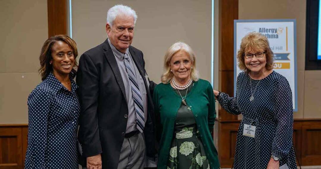 Four adults, three women and one man, stand together indoors, smiling at the camera. They are dressed semi-formally. A sign in the background mentions "Allergy" and "Asthma.