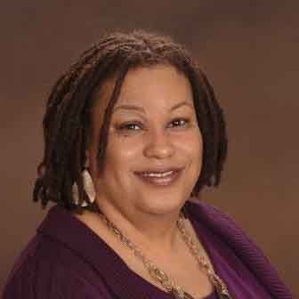 A person with short dreadlocks and wearing a purple top smiles warmly. They have hoop earrings and a necklace, posing against a plain brown background.