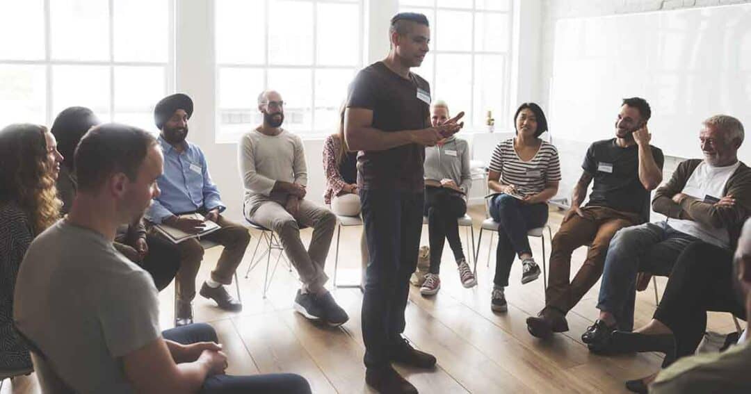A diverse group of people sit in a circle on chairs in a well-lit room, participating in a discussion. One person stands in the middle, speaking, while others listen attentively. The atmosphere appears casual and focused.