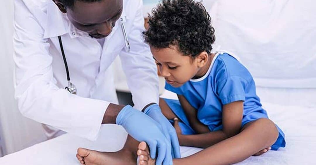 A doctor wearing gloves examines the foot of a young child sitting on a medical bed. The child is in a blue gown and seems focused on the examination. The setting appears to be a clinical environment.