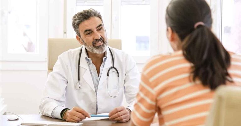 A male doctor with short gray hair and a beard, wearing a white coat and stethoscope, sits at a desk speaking with a female patient in a striped shirt. They are in a bright office with large windows in the background.