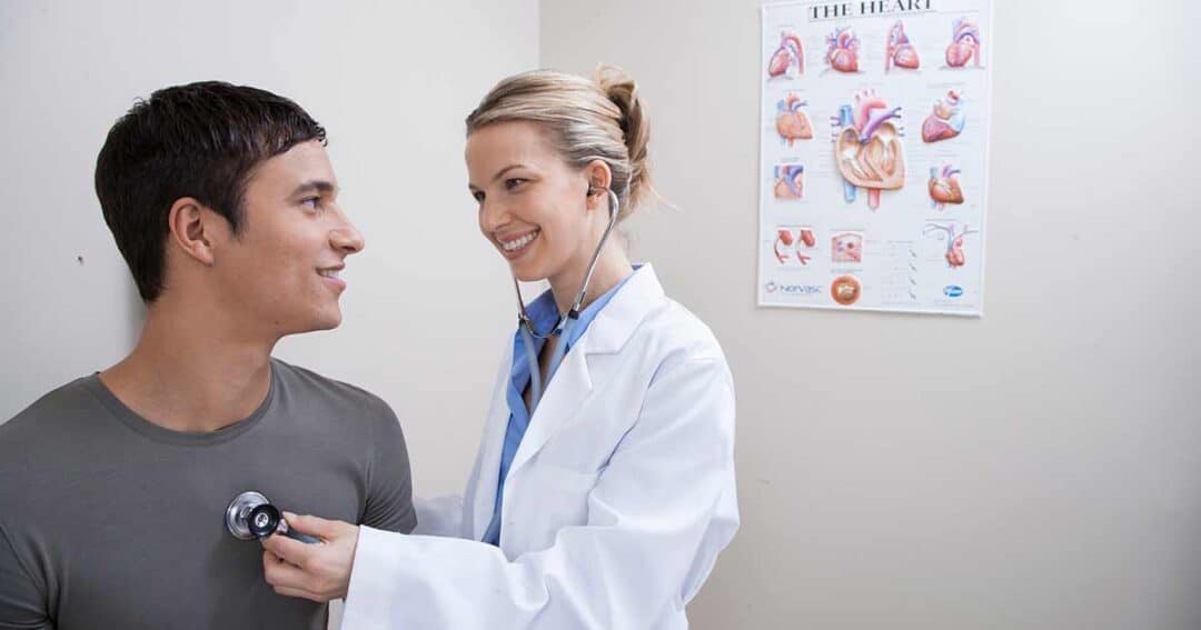 A doctor in a white coat uses a stethoscope to listen to the heartbeat of a patient sitting on an examination table. They are smiling at each other. A medical poster of the human heart is visible on the wall in the background.