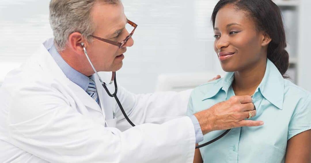 A doctor wearing a white coat and glasses uses a stethoscope to listen to the heartbeat of a smiling woman sitting in an examination room. The woman is wearing a light blue shirt.