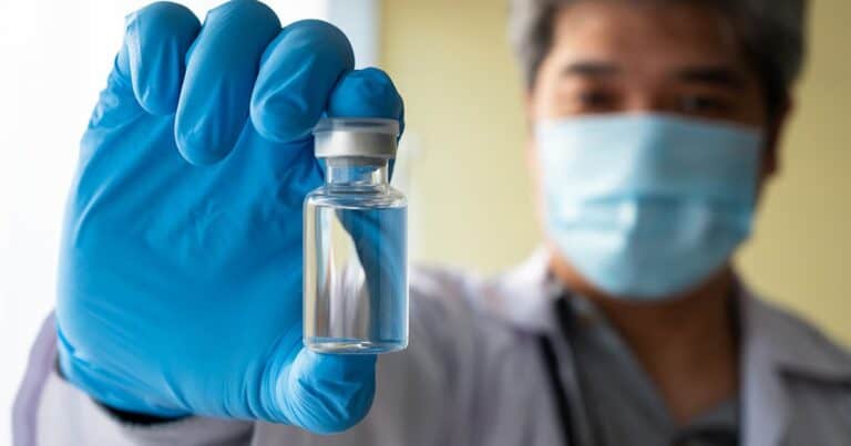 A healthcare worker wearing a mask and gloves holds up a clear vial, partially filled with liquid, in focus. The person is slightly out of focus in the background.