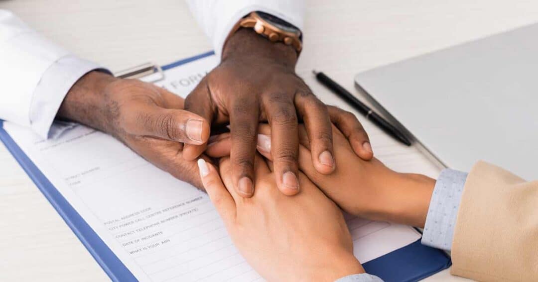 Two people are sitting at a table. One person's hands, dressed in a white coat, are gently resting on another's hands, offering reassurance. A clipboard with a form detailing health insurance options, alongside a pen and laptop, is visible on the table.