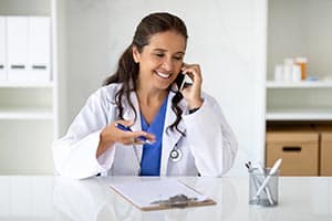 A smiling woman in a white lab coat sits at a desk, talking on a phone. She holds a pen and gestures with her hand. A stethoscope hangs around her neck. The background features shelves and office supplies.