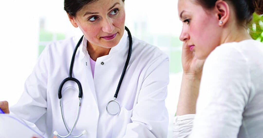 A doctor with a stethoscope talks to a concerned patient. Both women are engaged in conversation, with the doctor explaining something serious. The setting appears to be a medical office with bright lighting.