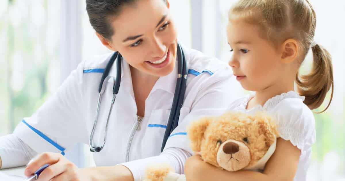 A smiling doctor with a stethoscope talks to a young girl holding a teddy bear. The doctor is wearing a white coat, and they both seem engaged in a positive interaction.