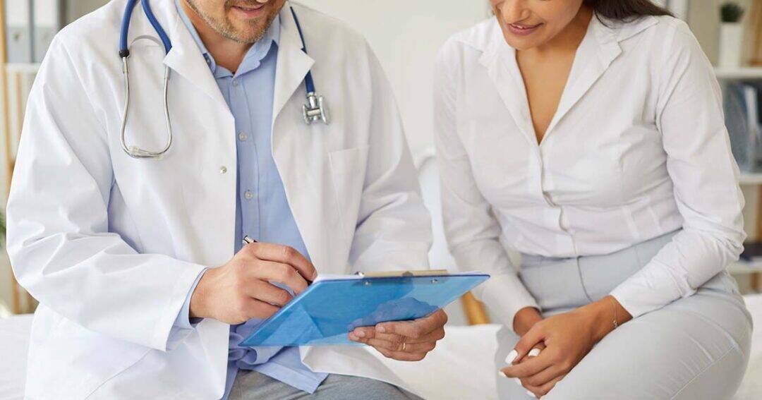 A doctor in a white coat reviews notes on a clipboard while sitting next to a patient, both wearing white shirts, in a medical office setting.