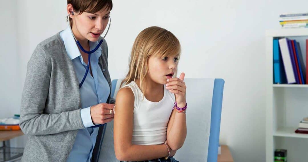 A doctor listens to a young girl's breathing with a stethoscope. The girl is sitting on an examination table, wearing a white tank top and jeans, and appears to be coughing. The room is bright and contains shelves and a desk.