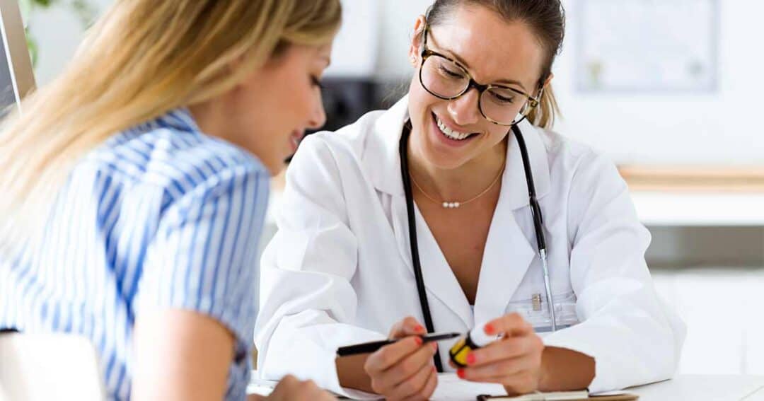 A doctor in a white coat and glasses smiles while showing a medication bottle to a patient with long blonde hair. The patient wears a blue and white striped shirt. Both are seated at a table in a bright office.