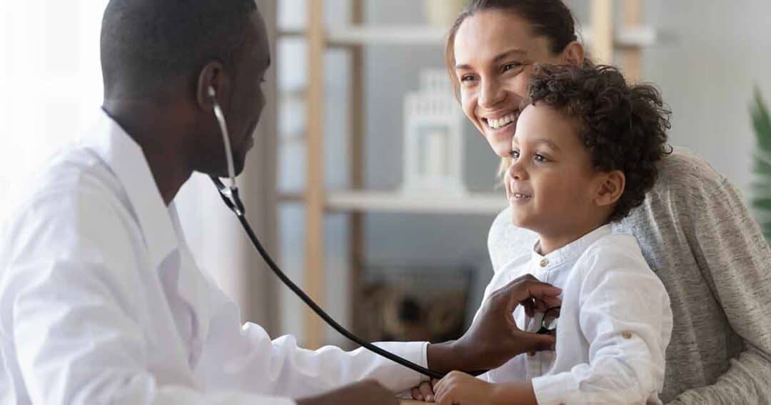 A doctor uses a stethoscope to check a young boy's heartbeat as he sits on his caregiver's lap. The caregiver smiles warmly, and the doctor is attentive. The setting is a well-lit, calm medical office.