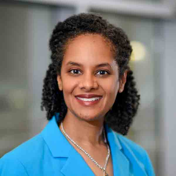 Black People Like Me Virtual Conference Series 1 A woman with curly hair wearing a blue blazer and a silver necklace smiles at the camera, with a blurred indoor background.