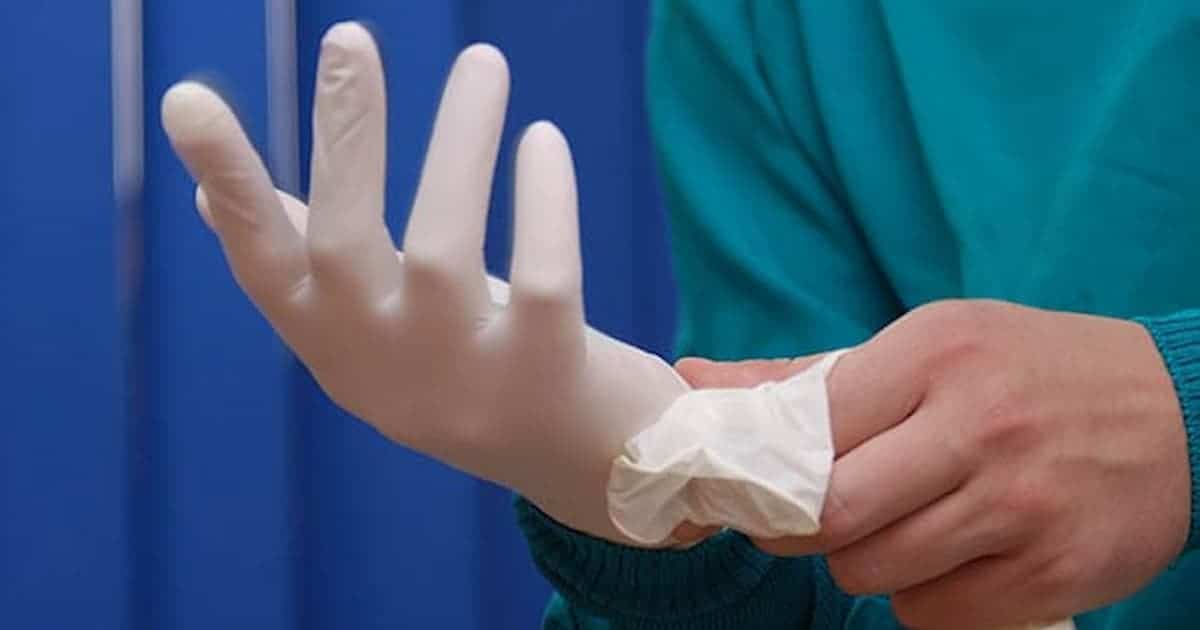 A person wearing a teal medical uniform is putting on a white latex glove, with a blue curtain visible in the background.