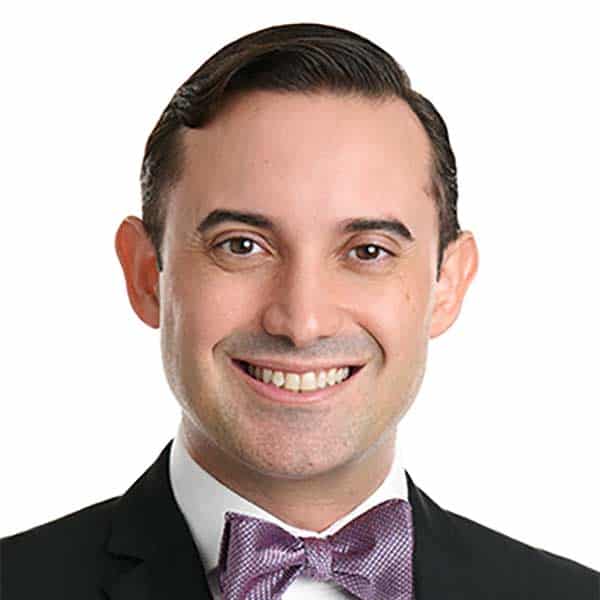 A man wearing a dark suit, white shirt, and a light purple bow tie is smiling at the camera against a plain white background.