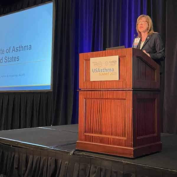 A woman stands at a podium with a microphone, presenting at the USAsthma Summit 2023. A large screen next to her displays a slide titled "The State of Asthma in the United States." The setting is a conference room with dark curtains.