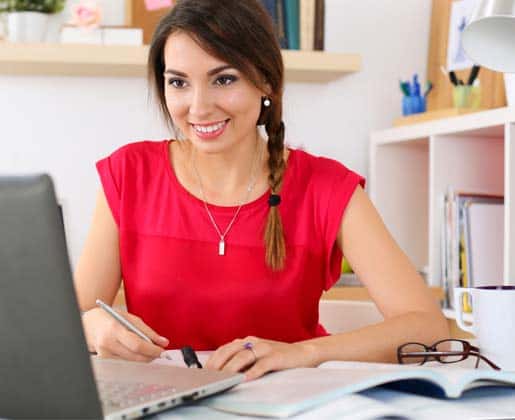Planning for School with Food Allergies 7 A woman with a braid and a red top sits at a desk, smiling while writing in a notebook. A laptop is open in front of her, with books, glasses, and a coffee mug nearby. The background has shelves with various office supplies.