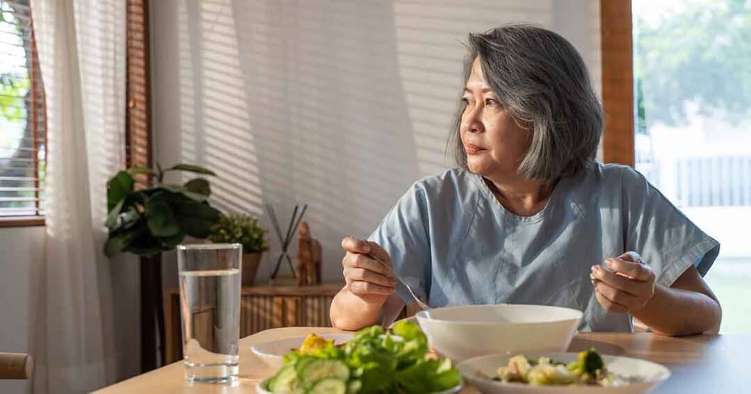 What Can You Do If You Can’t Afford Allergen-Free Foods? 8 A woman with gray hair sits at a dining table, looking thoughtfully out the window. There are plates of salad and a bowl in front of her. A glass of water is on the table, and sunlight filters through the window blinds.