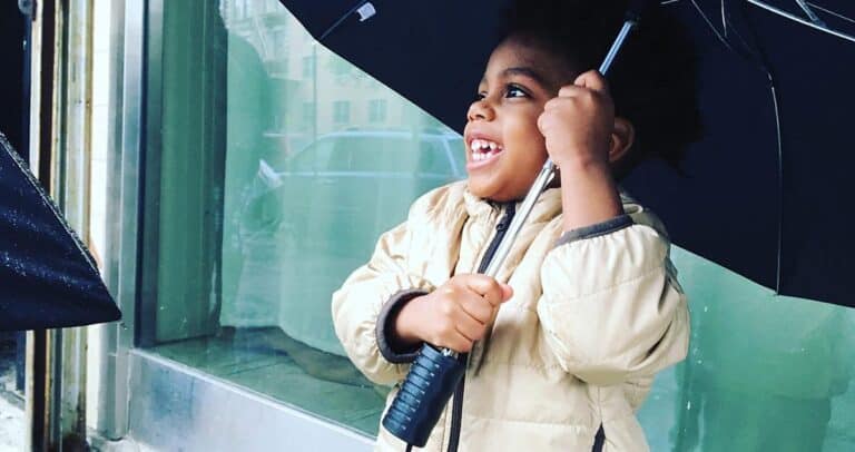 A young child in a beige jacket is joyfully holding an open black umbrella. They are smiling widely, standing outdoors in front of a glass window.