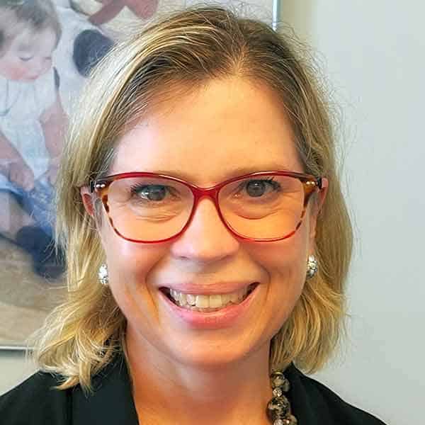 Person with shoulder-length blonde hair, wearing red glasses and a black top, smiling at the camera. A blurred painting is visible in the background.