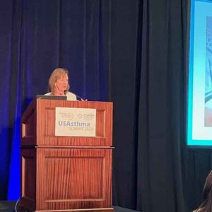 A person stands at a wooden podium with a sign that reads "US Asthma Summit 2023." They are speaking into a microphone on a stage with a dark curtain and a screen displaying a partial image on the right.