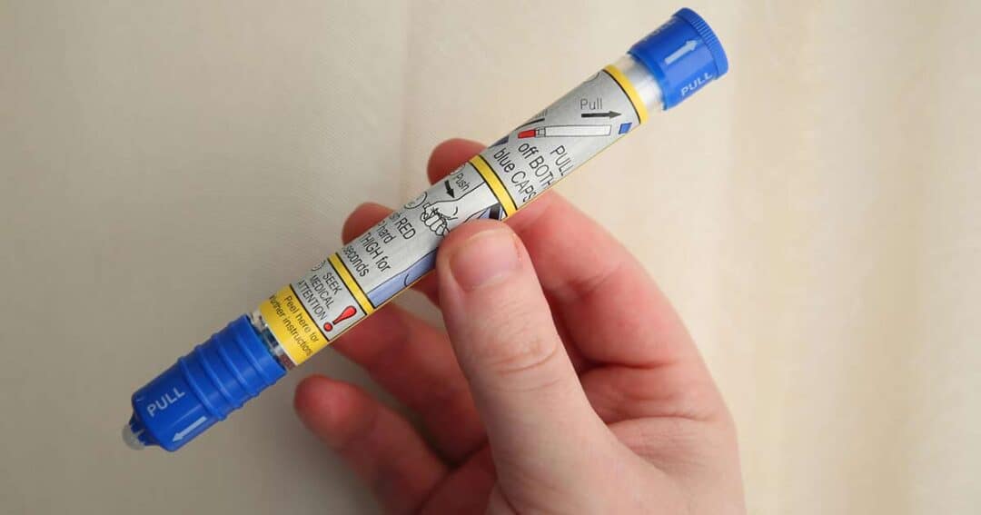A hand holding an epinephrine auto-injector pen with a blue cap and detailed instructions on its label, set against a beige background.