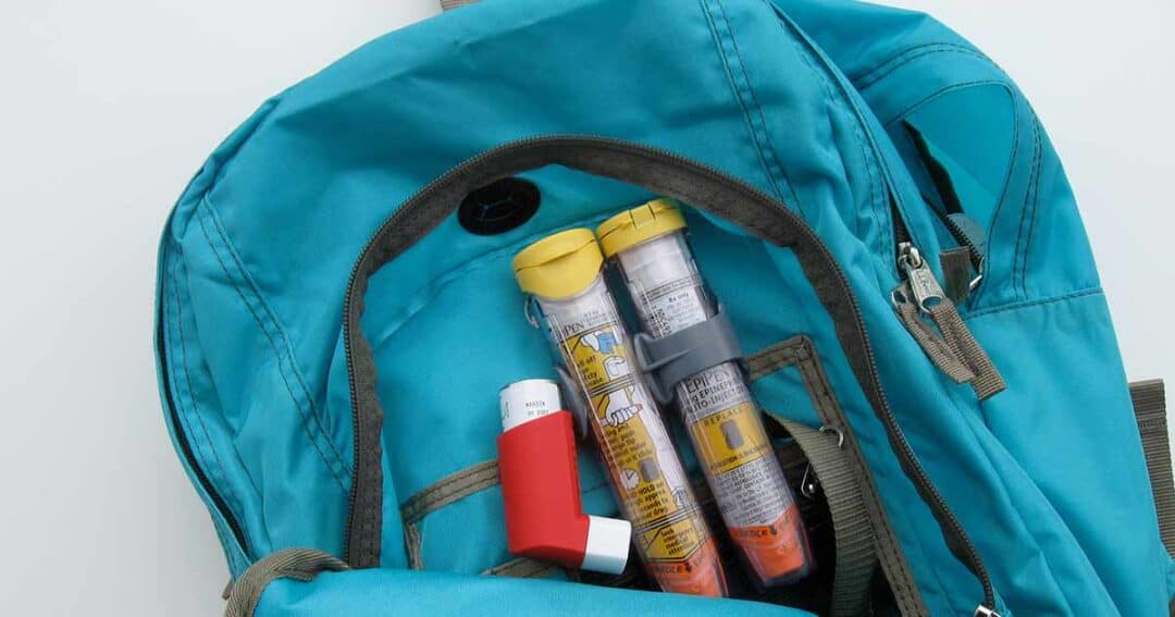 A blue backpack with an open pocket reveals three medical items: a red and white inhaler and two epinephrine auto-injectors with yellow caps.