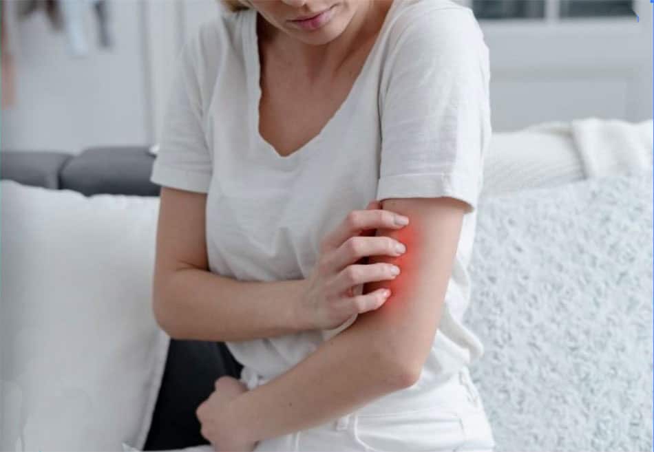 A person wearing a white shirt is sitting on a couch, scratching a red, irritated patch on their arm. The room appears to be softly lit, with a white blanket draped on the couch.