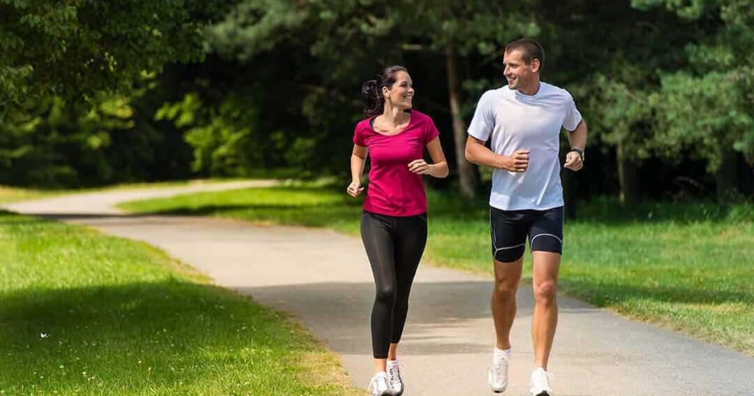 A man and a woman are jogging together on a paved path in a park. They are smiling at each other, surrounded by green grass and trees on a sunny day.