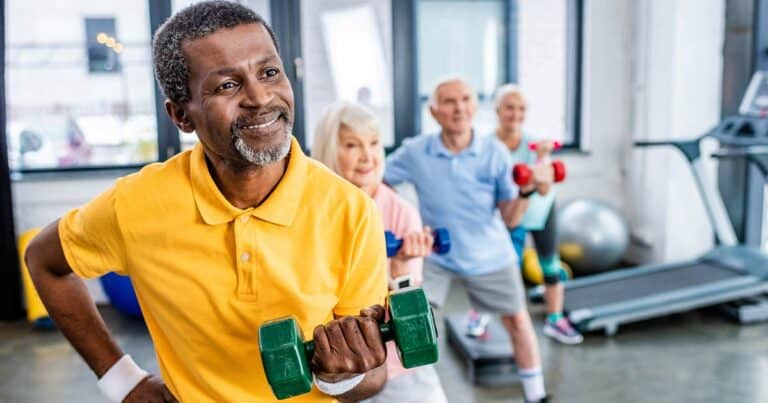 In a lively gym, a group of exercising seniors brings energy and joy. A man in a yellow shirt beams as he lifts a green dumbbell while three others lift weights. Nearby, a treadmill and exercise balls await their turn to contribute to the vibrant scene.