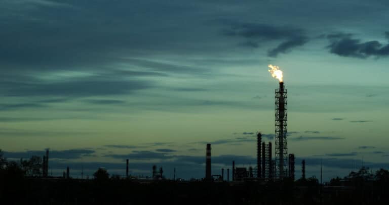A silhouetted industrial refinery emits a bright flame from a tall stack against a dusky, overcast sky. Dark outlines of trees and structures are visible in the foreground.