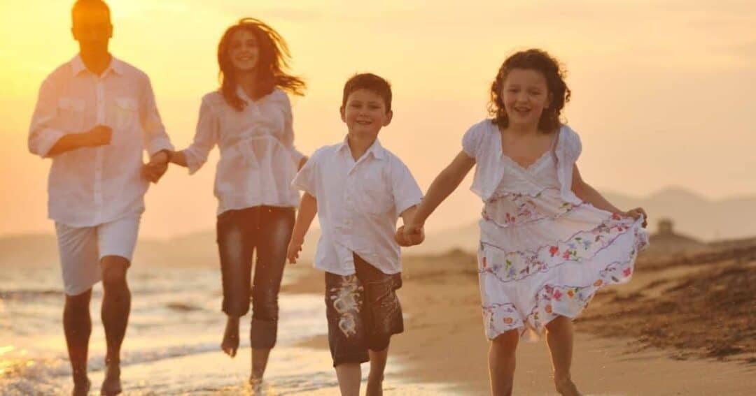 A family of four walks along a sandy beach at sunset. The parents, dressed in white, hold hands with their two children, a boy and a girl, who are smiling and dressed in summery outfits. The sun casts a warm glow over the scene.
