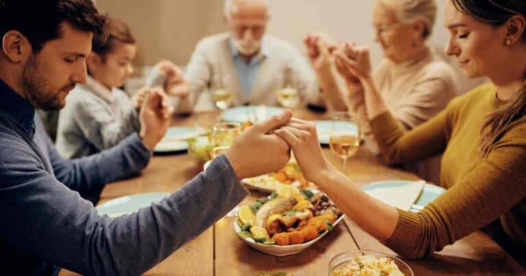 Family gathered around a dinner table, holding hands and praying. The table is set with plates of food and glasses of water and wine. Four adults, including an elderly couple, and a child are visible.
