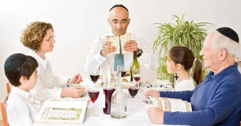 A family of five sits at a table during a Passover Seder. The man in the center holds matzah, while others hold books. The table is set with wine and festive dishes, and a tall plant is visible in the background.