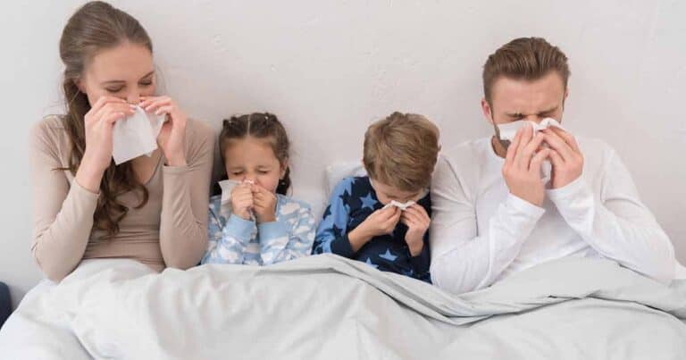 A family of four, two adults and two children, sit on a bed under a light grey blanket. All are wearing pajamas and blowing their noses into tissues, suggesting they have colds. The background is a plain white wall.