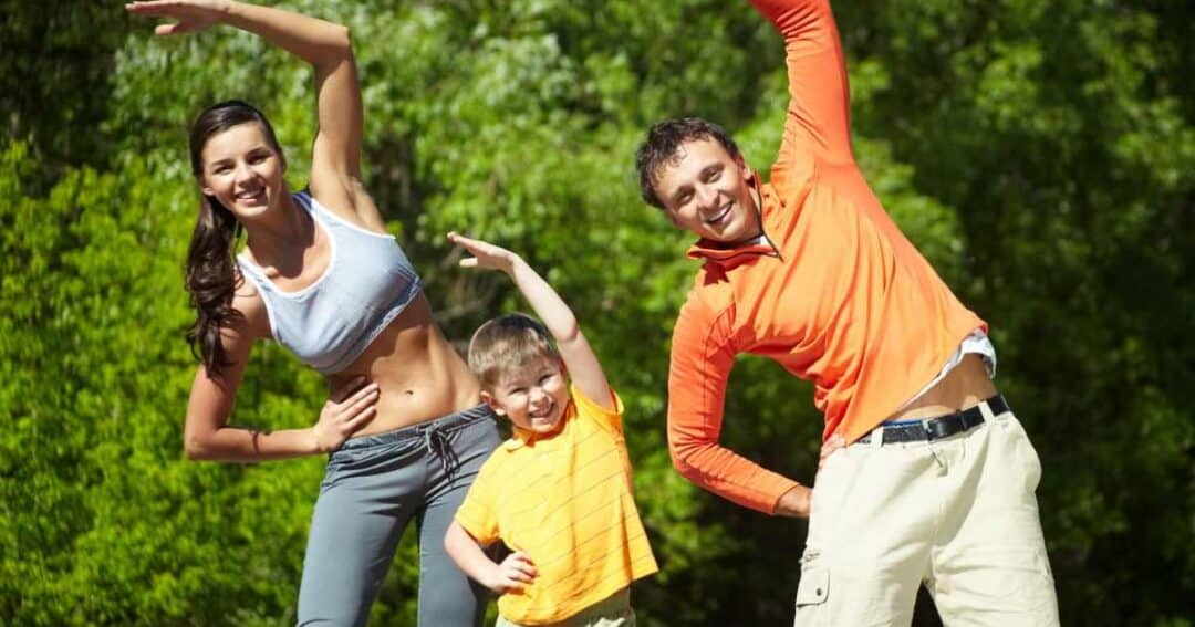 A family of three, a woman, a man, and a boy, are outdoors doing side stretches. They are smiling, wearing casual sportswear, and surrounded by greenery. The sun is shining brightly.