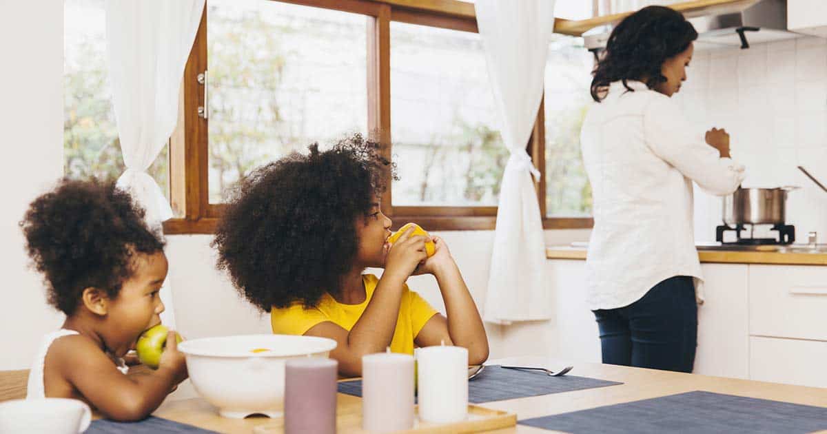 a mother is cooking while her children snack on fruit at the kitchen table