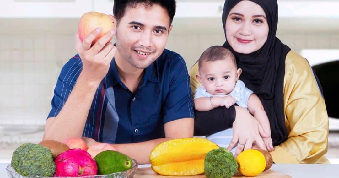 A family of three in a kitchen, with a man holding an apple and a woman holding a baby. A table in front displays fruits and vegetables, including broccoli, papaya, lemons, avocado, and a dragon fruit.