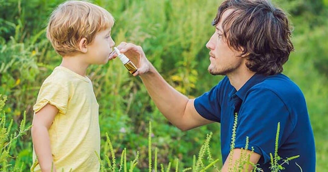 Grass Allergy 1 A man kneels on the grass while giving a young child a drink from a small cup. The child is wearing a yellow shirt, and the man is in a blue shirt. They are surrounded by greenery, with plants and grass in the background.