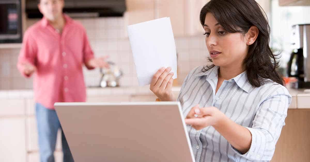 A woman sits at a table with a laptop, holding a piece of paper and looking concerned, perhaps considering the implications of food allergies. In the background, a man in a pink shirt stands with arms outstretched, appearing to speak about mental health support. They are in a kitchen setting.