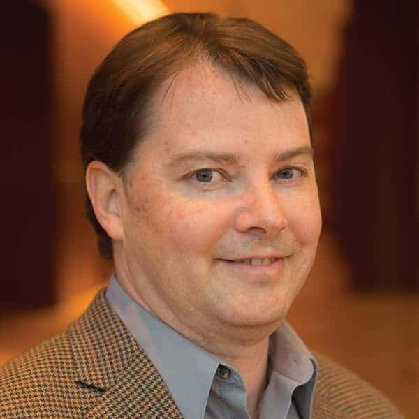A man with short brown hair smiles slightly while wearing a checked blazer and a light gray shirt. The background is softly blurred with warm lighting.