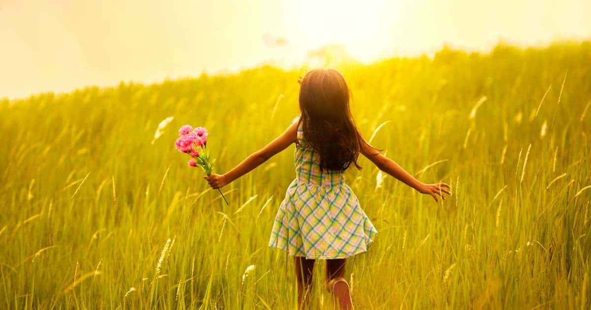 A young girl in a plaid dress runs through a sunny field holding a bouquet of colorful flowers. The tall grass and warm sunlight create a serene and joyful atmosphere.