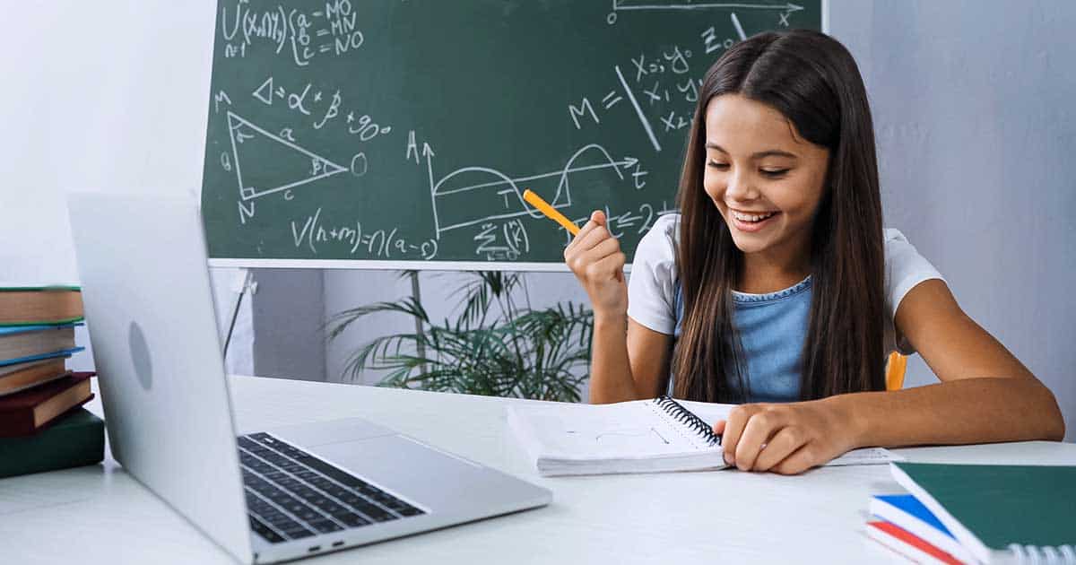 A girl is sitting at a desk with a laptop, notebooks, and her food allergy journal, focusing on learning food allergy tips. She is holding a pencil and smiling. In the background, a chalkboard brims with mathematical formulas and diagrams.