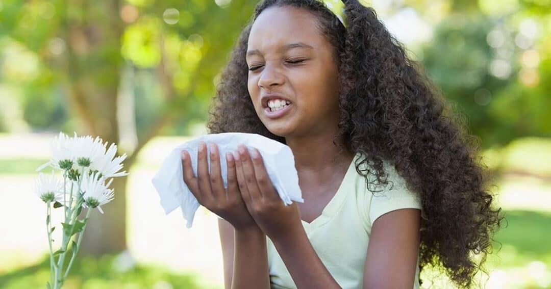 A young girl with curly hair sneezes into a tissue while sitting outdoors. She appears to be reacting to white flowers nearby, likely due to allergies. The background is a vibrant green garden.